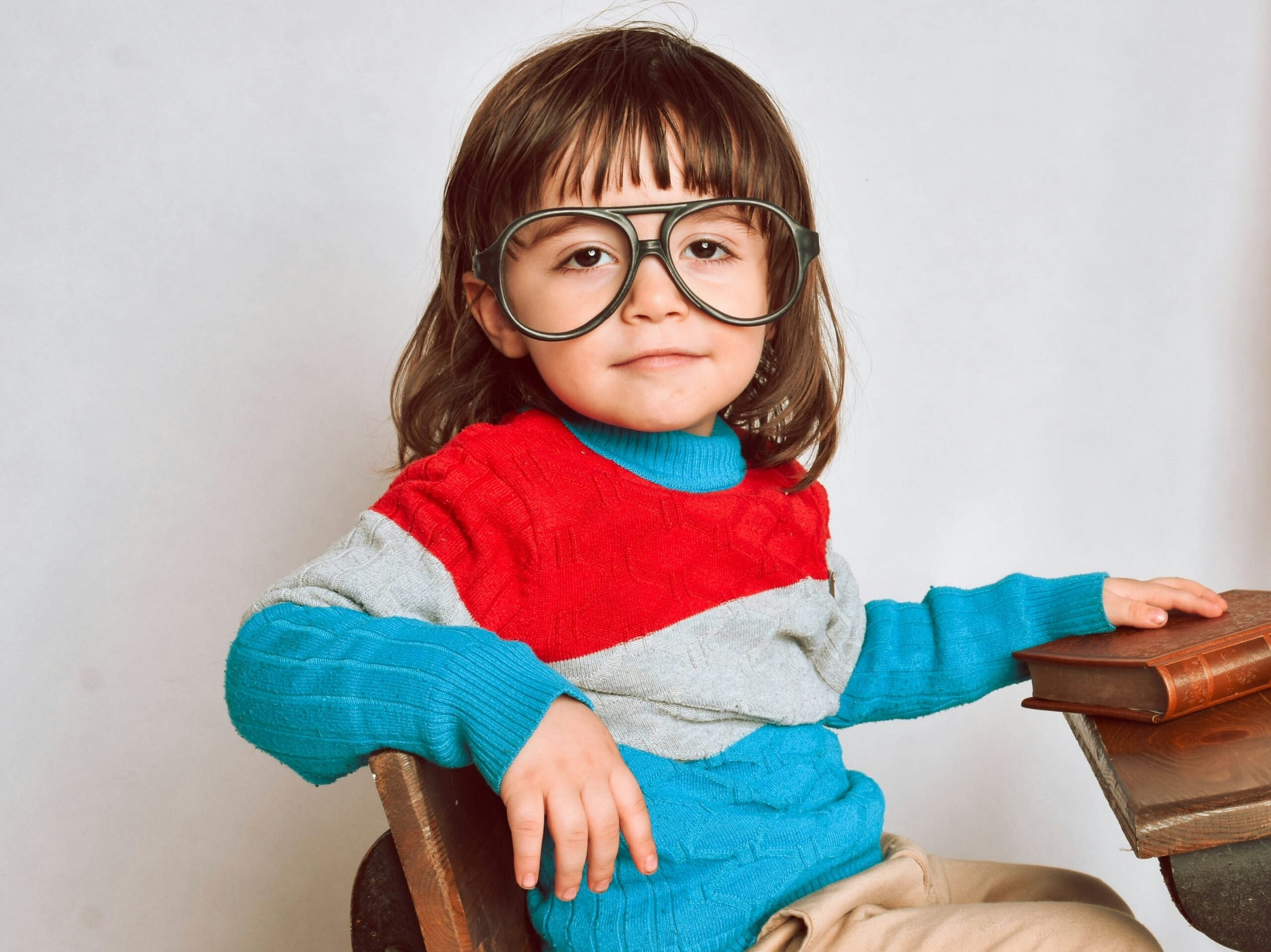 A child with bangs, sitting on a chair and wearing eyeglasses